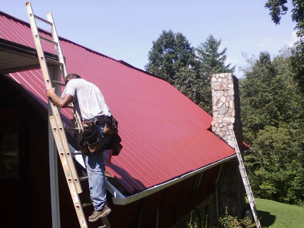 Roofer on a ladder beside a red metal roof and stone chimney — Soffit & Fascia Repair by Roofing Brothers in Centerbrook